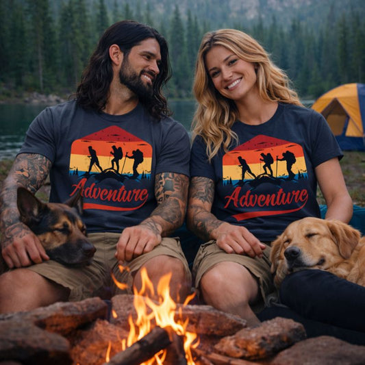 Two people and two dogs sitting by a campfire with 'Adventure' t-shirts in a forest setting.
