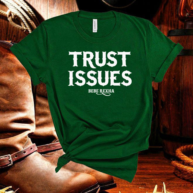 Green t-shirt with 'Trust Issues' text in white, worn over brown boots on a wooden floor.