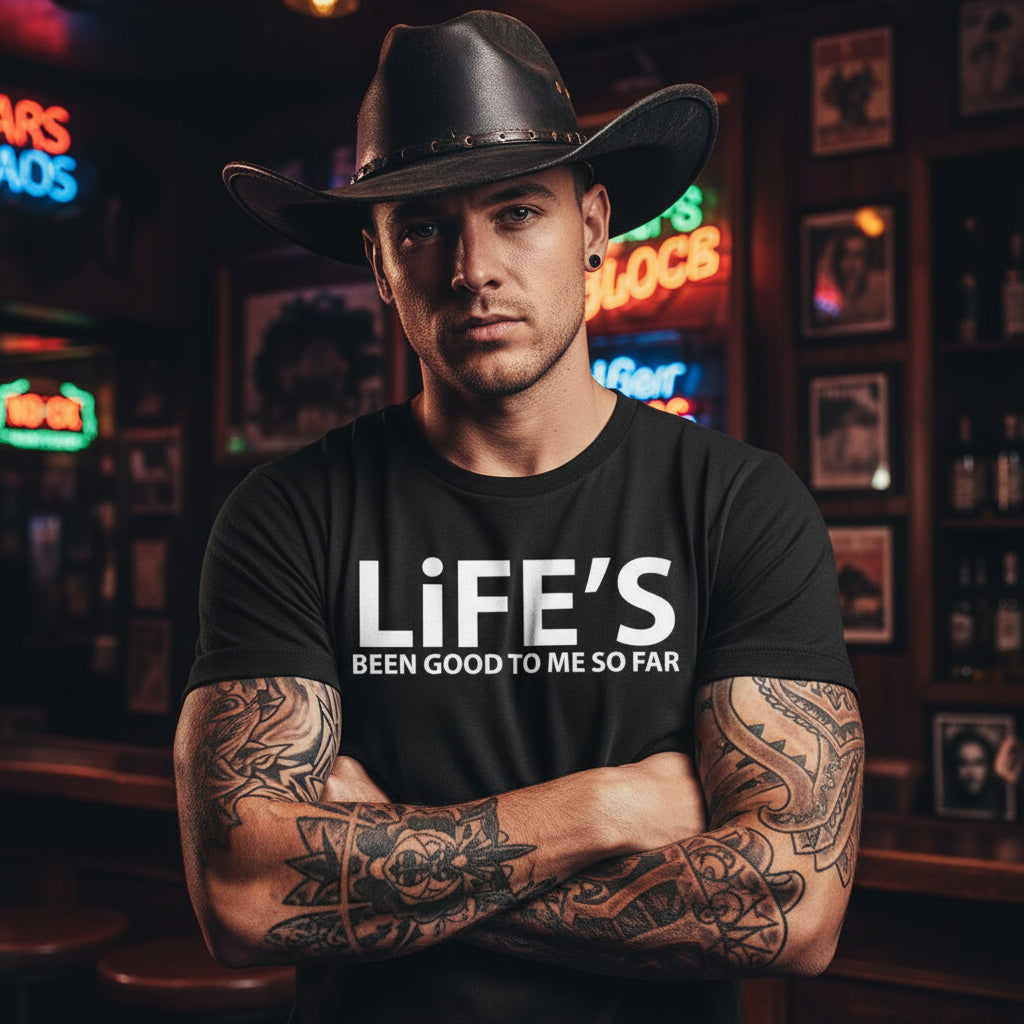 Black t-shirt with 'LiFE'S BEEN GOOD TO ME SO FAR' text against a dramatic sky and rustic building background