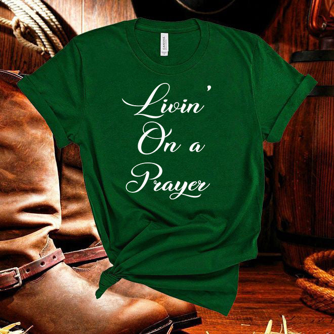 Green t-shirt with 'Livin' On a Prayer' text on a wooden background with cowboy boots and rope.