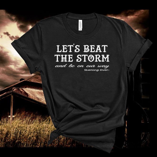 Black t-shirt with 'Let's beat the storm and be on our way starting over' text against a dramatic sky and rustic building background.