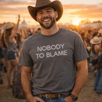 Man wearing a 'Nobody to Blame' t-shirt at a concert with a sunset in the background