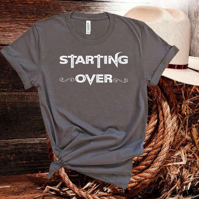 Gray t-shirt with 'STARTING OVER' text on a wooden surface with a hat and straw basket.