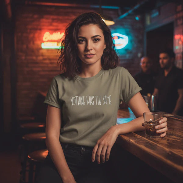 Woman in a bar wearing a t-shirt with 'Nothing Was the Same' text, holding a glass.