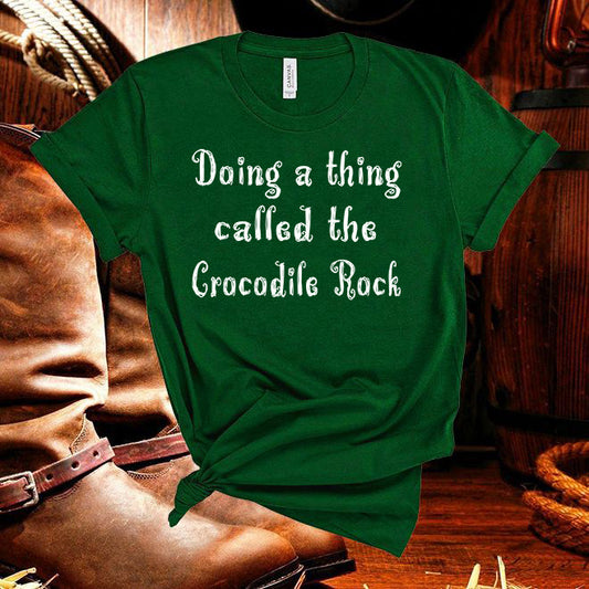 Green t-shirt with text 'Doing a thing called the Crocodile Rock' on a wooden background with cowboy boots.