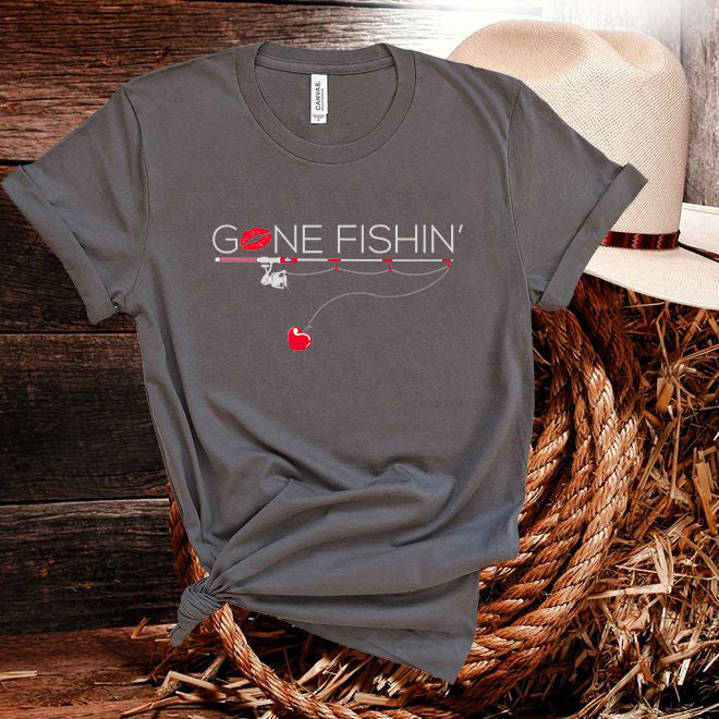 Gray t-shirt with 'Gone Fishin'' design on a wooden surface with a hat and basket.