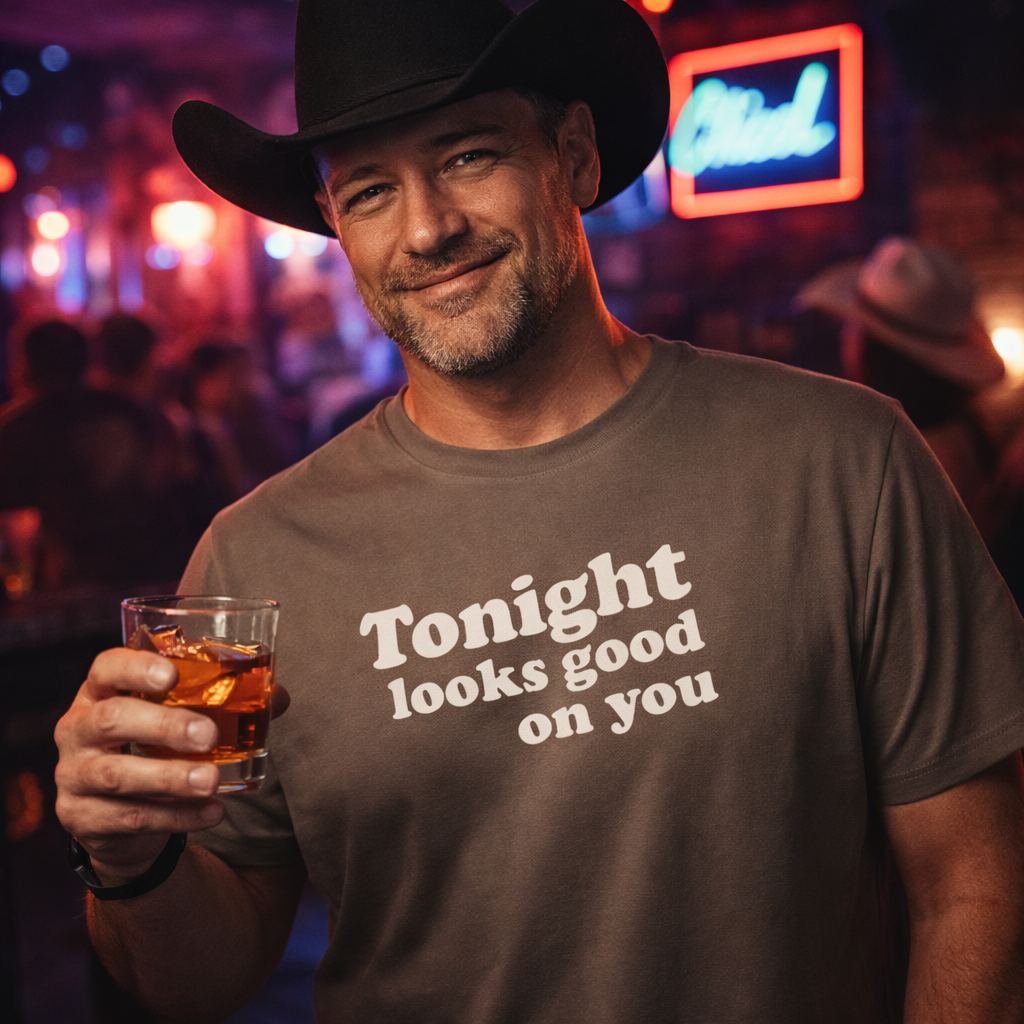 Man wearing a cowboy hat and 'Tonight looks good on you' shirt holding a drink in a bar setting.