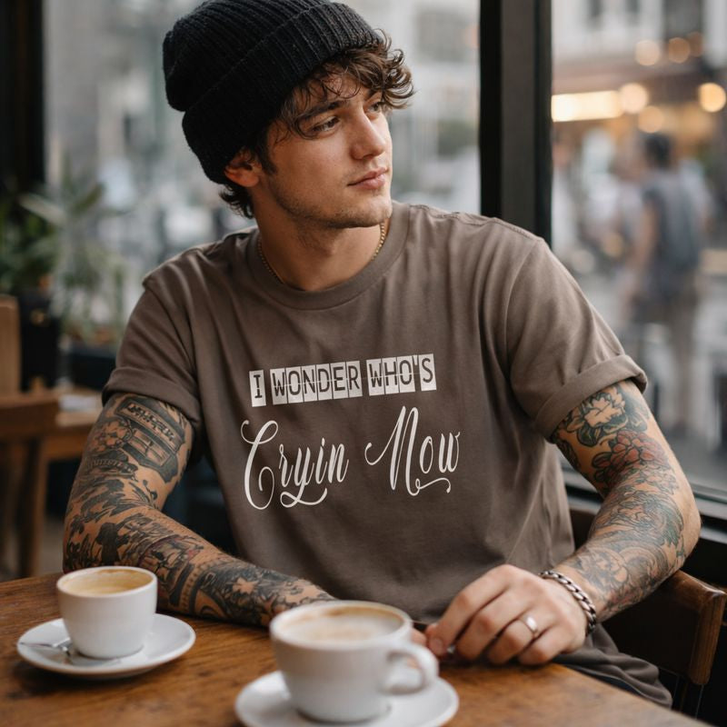 Man with tattoos wearing a shirt with text, sitting at a cafe with coffee.