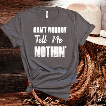 Gray t-shirt with text 'CAN'T NOBODY TELL ME NOTHIN'' on a wooden surface with a hat.