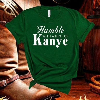 Green t-shirt with 'Humble with a Hint of Kanye' text on a wooden background with cowboy boots.