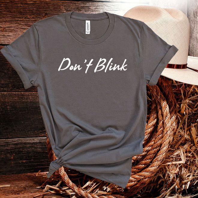 Gray t-shirt with 'Don't Blink' text on a wooden surface with a straw hat.