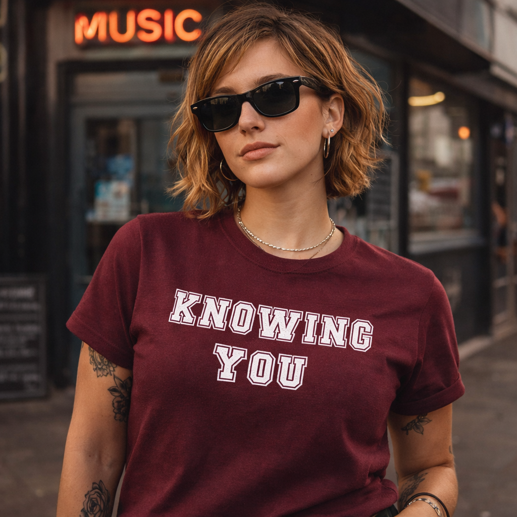 Person wearing a maroon t-shirt with 'KNOWING YOU' text in an urban setting.