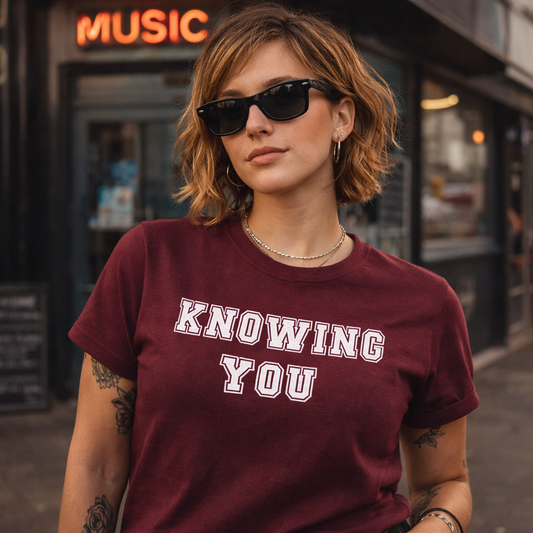 Person wearing a maroon t-shirt with 'KNOWING YOU' text in an urban setting.