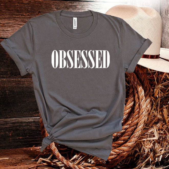 Gray t-shirt with 'OBSESSED' text on a wooden surface with a hat.